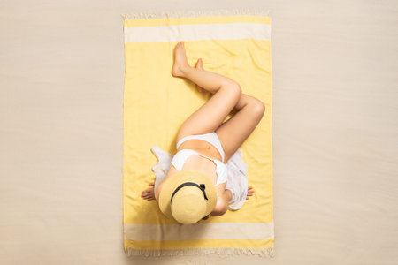Alone woman in straw hat and bikini sitting and sunbathes on yellow beach towel. Female relaxation...