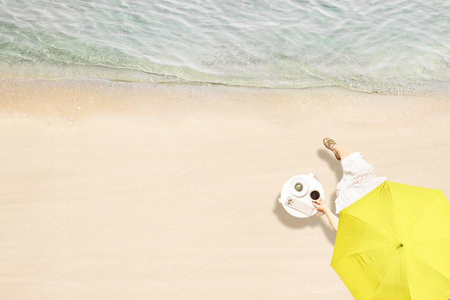 Woman drinking hot coffee with cake sitting at table under beach umbrella near sea coastline of sand beach. Summer vacation. Outdoors cafe. Aerial viewの写真素材