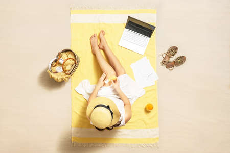 Businesswoman freelancer applying sunscreen on skin sitting on the beach towel of sand beach and working on laptop with graphics and charts. Summer. Top viewの写真素材