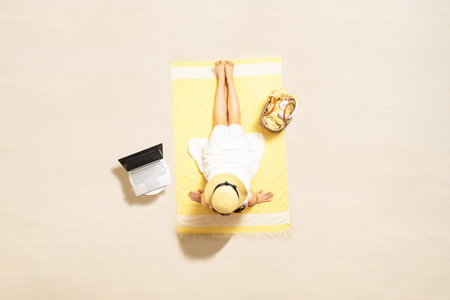Woman with laptop, bag and headphones sitting on the beach towel and enjoying summer vacation....