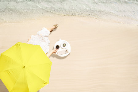 Woman drinking hot coffee with cake sitting at table under beach umbrella near sea coastline of sand...