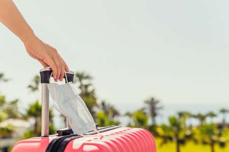 Woman with pink suitcase, protective mask standing on passengers ladder and getting out of airplane opposite sea coastline with palm trees.Tourism conceptの写真素材