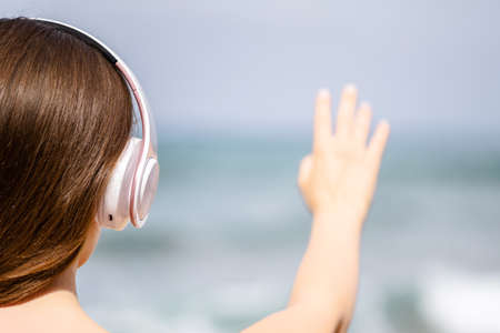 Happy woman in headphones listen music waving hand to say hello and welcoming sea standing on the beach. Summer vacation. Back viewの写真素材