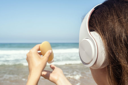 Woman in headphones listen music applying sunscreen on skin on a beach looking on the sea. Female...