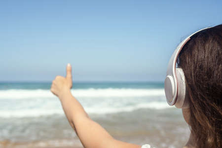Happy woman on a beach in headphones listen music, showing thumb up looking on the sea. Female relaxation at summer vacation. Back viewの写真素材
