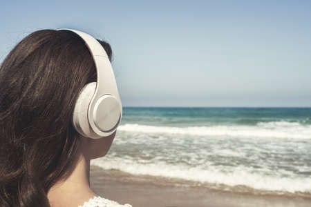 Back view of alone woman sitting on a beach in headphones listen music dreaming and looking on the sea. Female relaxation at summer vacation.の写真素材