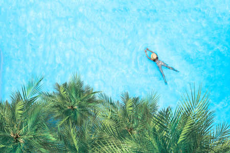 Young alone active woman in bikini and hat is swimming in swimming pool with palm trees. Aerial drone view from above. Tropical summer vacationの写真素材