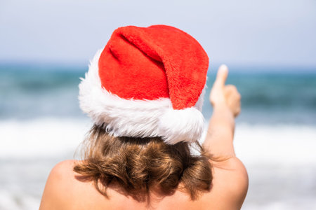 Christmas sea holiday. Back view of happy woman in santa hat showing thumb up and relaxing on...