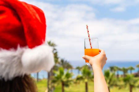 Woman hands in christmas gloves and Santa Claus hat relaxing and drinking cocktail on balÑony opposite palm trees beach and sea. New yearの写真素材