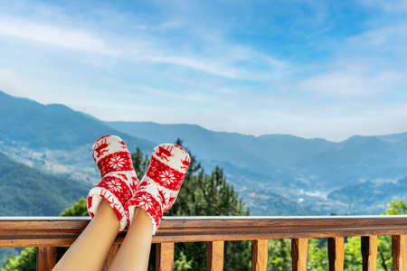 Woman in Christmas red socks, boots sits on a balcony in the mountains putting her feet on the wooden terrace railingの写真素材