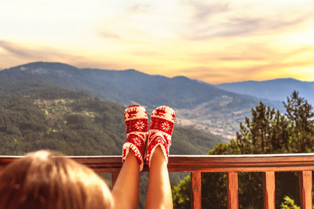 Woman in Christmas red socks, boots sits on a balcony in the mountains at sunsetの写真素材