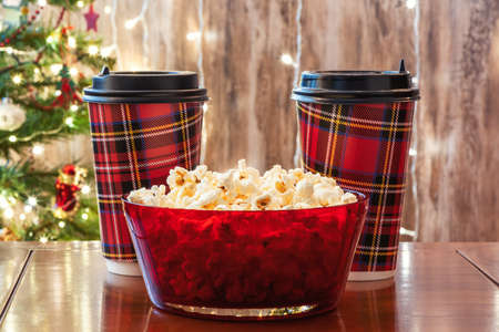 Christmas eve. Red bowl with popcorn and two paper cups of drinks, cola on the table near christmas tree. Home cinema. Cropped, close upの写真素材