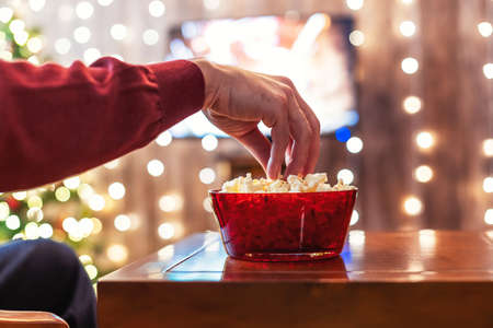 Christmas eve. Alone man watching tv and eating popcorn sitting near christmas tree. Home cinema. Cropped, close upの写真素材