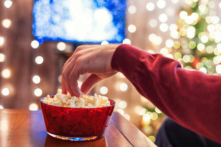 Christmas eve. Alone man watching tv and eating popcorn sitting near christmas tree. Home cinema. Cropped, close upの写真素材