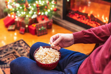 Christmas eve. Alone man watching tv and eating popcorn sitting near fireplace and Christmas tree. Home cinema. Cropped, close upの写真素材