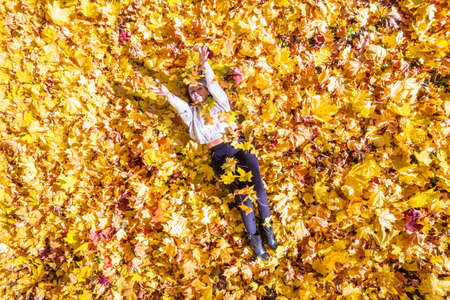 Top view. Beautiful happy young woman in red sweater lying on ground having fun, playing, toss up with autumn maple autumn leaves in park. Aerial, drone view. Indian summerの写真素材