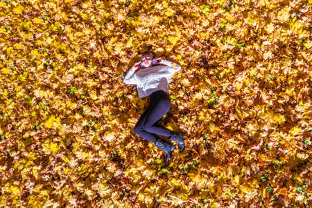 Top view. Beautiful happy young woman in headphones listening music lying and relaxing on the autumn maple leaves in the park. Aerial, drone viewの写真素材