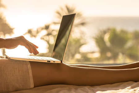 Woman with laptop sitting on the bed in bedroom opposite panoramic window with view on palm tree sea beach at sunsetの写真素材