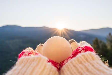 Female hands in warm knitted mittens holding a Christmas ball against the background of the mountainsの写真素材