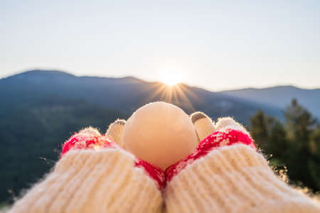 Female hands in warm knitted mittens holding a Christmas ball on the background of mountainsの写真素材