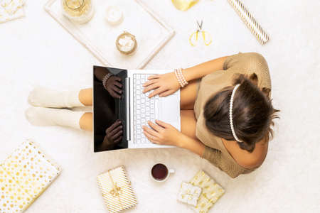 Christmas. Woman in sweater using laptop for searching gift ideas sitting on the white carpet among the many wrapped boxes in white gold wrapping paper. Top view. Conceptの写真素材