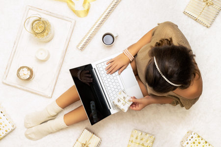 Christmas. Woman in sweater using laptop for searching gift ideas sitting on the white carpet among...