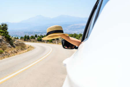 Woman driving a car holding straw hat from the open window. Trip on the serpentine road in the mountains. Summer vacationの写真素材