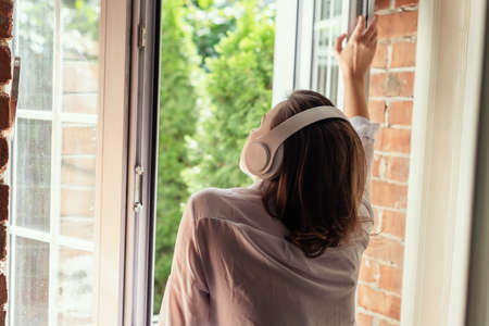 Young beautiful happy woman in headphones listening music opens window of country house and breathes fresh air in the early summer morning. Back view.の写真素材