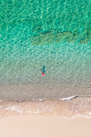 top, aerial view. Young beautiful woman in a hat and white bikini swimming in sea water on the sand...
