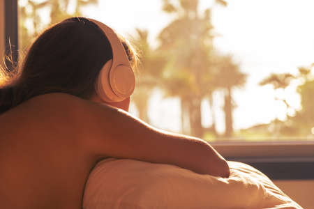 Silhouette of woman with headphones lying on pillow on the bed in bedroom opposite panoramic window with view on palm tree sea beach at sunset. Female relaxing listening music.の写真素材