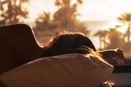 Silhouette of woman with headphones lying on pillow on the bed in bedroom opposite panoramic window with view on palm tree sea beach at sunset. Female relaxing listening music.の写真素材
