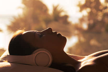 Silhouette of woman with headphones lying on pillow on the bed in bedroom opposite panoramic window with view on palm tree sea beach at sunset. Female relaxing listening music.の写真素材