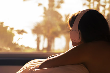 Silhouette of woman with headphones lying on the sunbed on terrace and looking at palm trees sea beach at sunset. Female relaxing listening music. back view.の写真素材