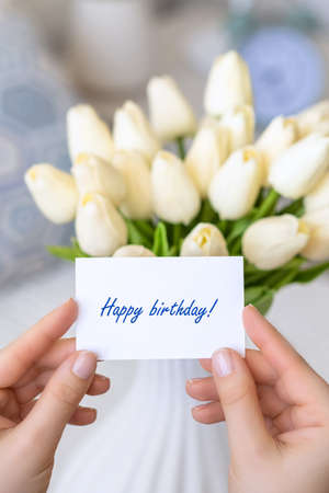 Woman pulling greeting card with Happy birthday words from bouquet of white tulips flowers in a white vase on the table.の写真素材