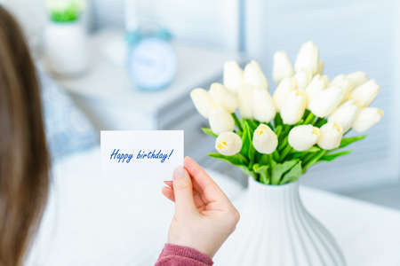 Woman pulling greeting card with Happy birthday words from bouquet of white tulips flowers in a white vase on the table.の写真素材