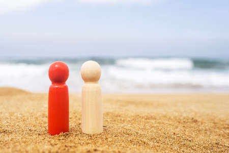 Wooden two figures of different color of people red, dark and white on the sand of beach with sea view. Concept of happy friends on holiday. International, different race.の写真素材