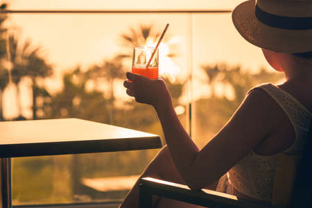 Rear view of woman with headphones and cup of tea sitting on chair near panoramic window and looking at palm trees sea beach at sunset. Female relaxing listening music.の写真素材