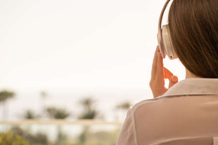 Rear view of woman with headphones and cup of coffee looking on palm trees sea beach at sunrise morning. Female relaxing listening music.の写真素材