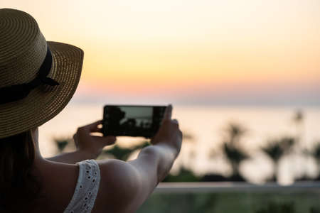 Rear view of woman looking at phone sitting on armchair near panoramic window opposite palm trees sea beach at sunset.の写真素材