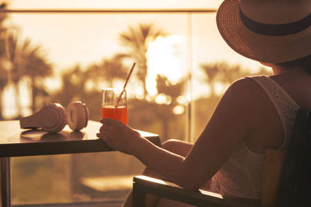 Rear view of woman looking at phone sitting on armchair near panoramic window opposite palm trees sea beach at sunset.の写真素材