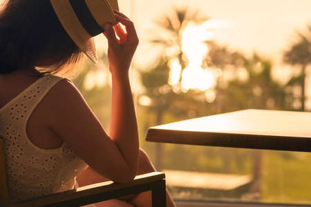Rear view of woman applying cream sitting on armchair near panoramic window and looking at palm trees sea beach at sunset.の写真素材