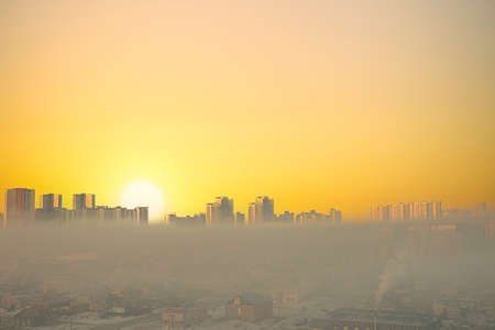 Winter, snow town buildings, skyscrapers on horizon with the huge setting sun. Village at early morning fog and orange pastel sky at sunsetの写真素材