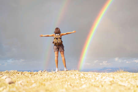 A tourist traveler girl with a backpack standing on the top of the mountain and looking at the rainbow on the blue sky background.の写真素材