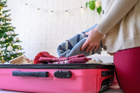 Christmas time. Woman putting knitted winter clothing in a suitcase in the room decorated christmas...