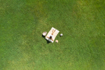 Woman in a white dress and hat, sitting on the plaid on the green grass having a picnic with food basket. Top view, drone, aerial viewの写真素材