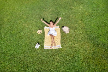 Woman in a white dress and hat, sitting on the plaid on the green grass having a picnic with food basket. Top view, drone, aerial viewの写真素材