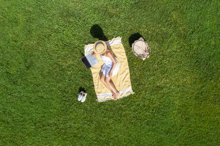 Woman in a white dress and hat, sitting on the plaid on the green grass having a picnic with food basket. Top view, drone, aerial viewの写真素材
