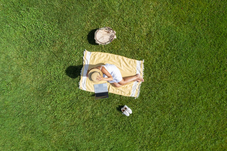 Woman in a white dress and hat, sitting on the plaid on the green grass having a picnic with food basket. Top view, drone, aerial viewの写真素材