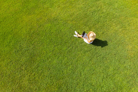 Woman in a white dress, hat with laptop, sitting on the green grass at summer day. Top view, drone, aerial viewの写真素材