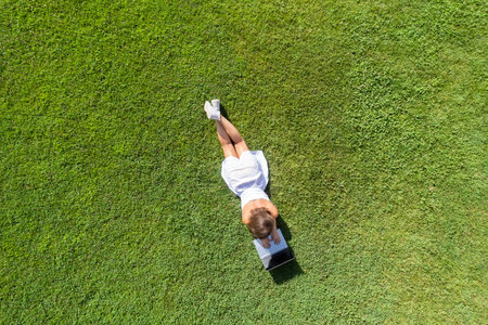 Woman in a white dress, hat with laptop, lying on the green grass at summer day. Top view, drone, aerial viewの写真素材